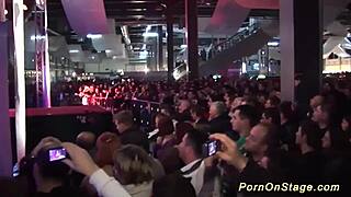 Two european blondes undress and kiss on public stage, reaching orgasm with voyeur watching.